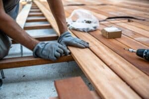 A worker wearing gloves is carefully aligning wooden planks while building a deck, with tools and materials like a drill and screws visible on the surface.